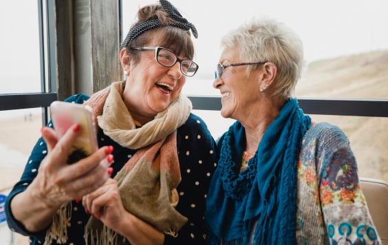 Two older ladies looking at one another laughing. One of them is showing the other something on her phone.