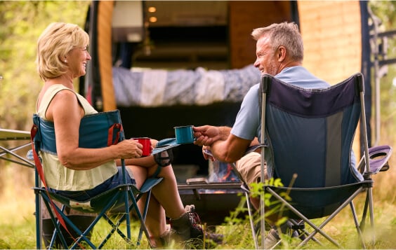Rear View Of Senior Couple Camping In Countryside With RV Drinking Coffee By Outdoor Fire