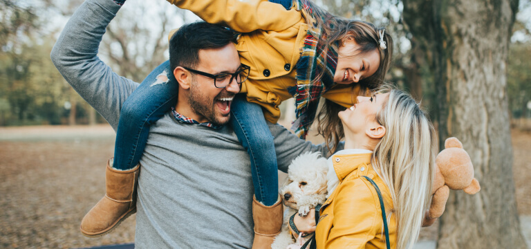 Young family with a dad carrying a young daughter on his shoulders as mother smiles at them 