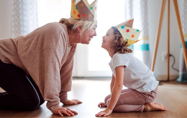Grandmother and her young granddaughter sat on their knees, smiling at one another whilst wearing party hats