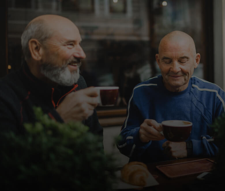 Two older men sitting outside a cafe drinking hot drinks