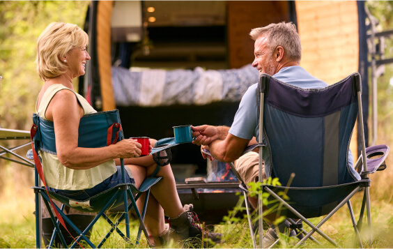 Rear View Of Senior Couple Camping In Countryside With RV Drinking Coffee By Outdoor Fire