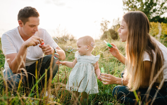 Daughter and parents on green and yellow grass and blowing soap bubbles in sunlight summer park. 