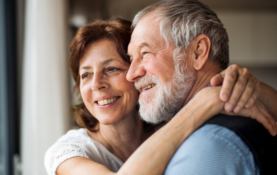 A senior couple indoors at home, looking out of window.