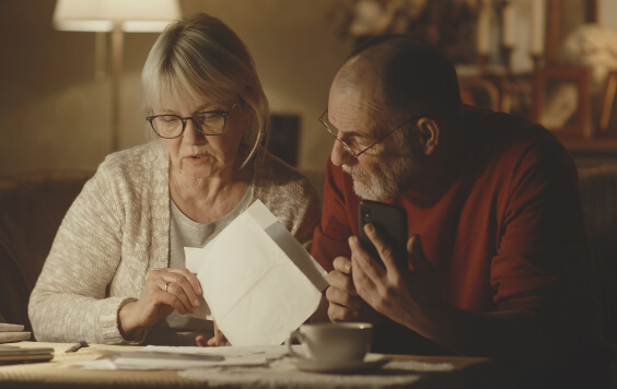 Preoccupied senior husband and wife analysing papers and making calculations while checking utility bills in dim living room at home 