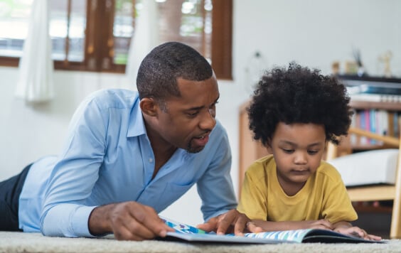 Cute son and his handsome father are reading a book while lying on floor in living room floor.