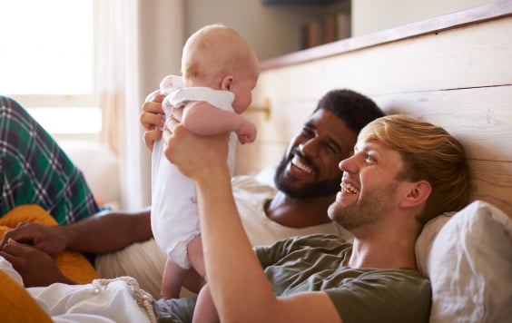 Loving Male Same Sex Couple Cuddling Baby Daughter In Bedroom At Home Together