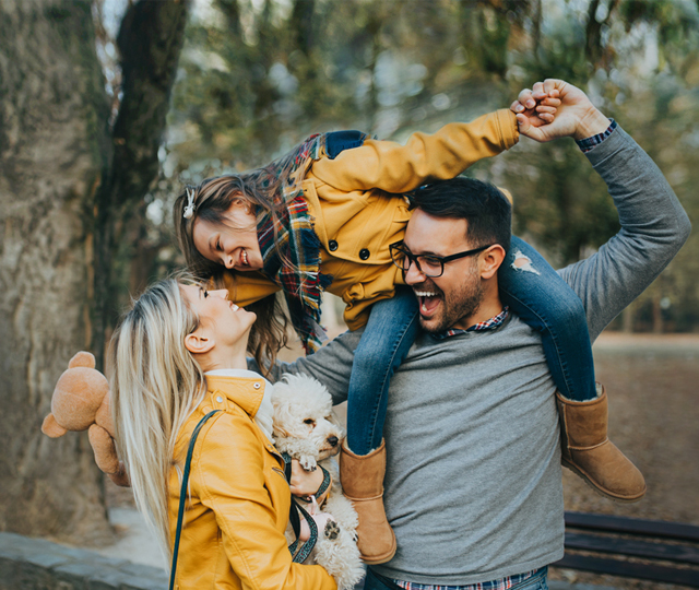Child atop happy father's shoulders, leaning over to kiss mother who's holding a small dog