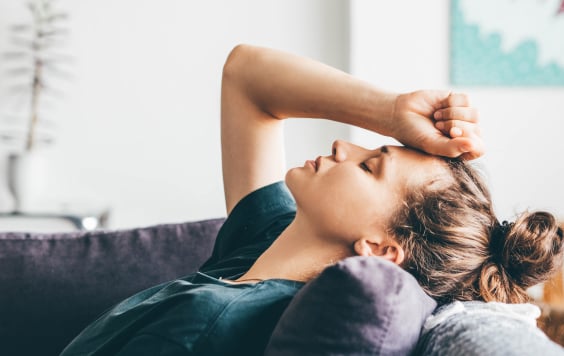 Exhausted woman laying back on sofa with their eyes closed and fist against their forehead